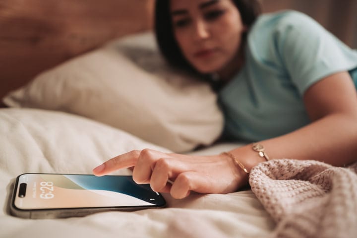 Young Woman's Hand Reaching For Her Smartphone On A White Duvet
