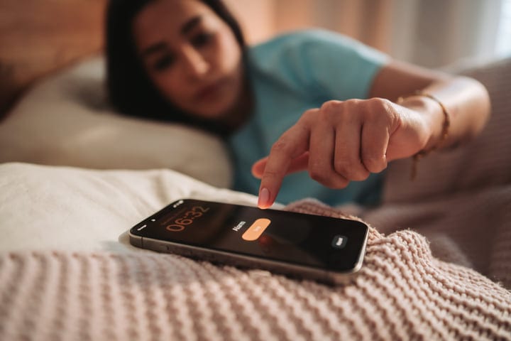 Young Woman Waking Up And Silencing Her Smartphone Alarm In Bed
