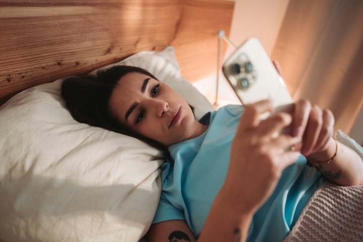 Young Woman Lying In Bed Browsing Her Smartphone In The Morning