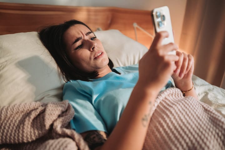 Young Woman Lying In Bed Frowning While Looking At Her Smartphone