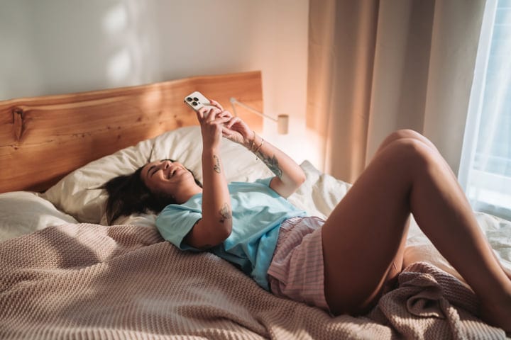 Smiling Woman Checking Her Phone In Bed In The Morning Light