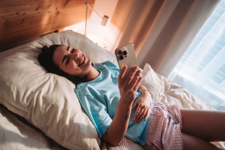 Young Woman Laughing While Relaxing in Bed and Browsing Her Smartphone