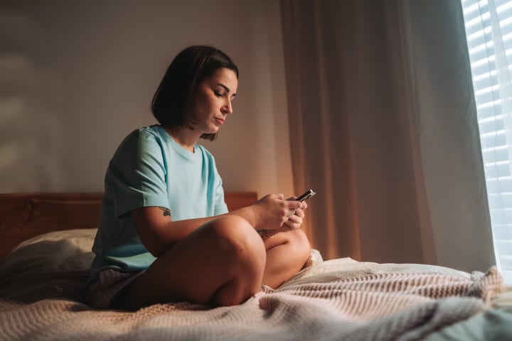 Young Woman Sits Cross-Legged On Bed Checking Smartphone In Morning Light