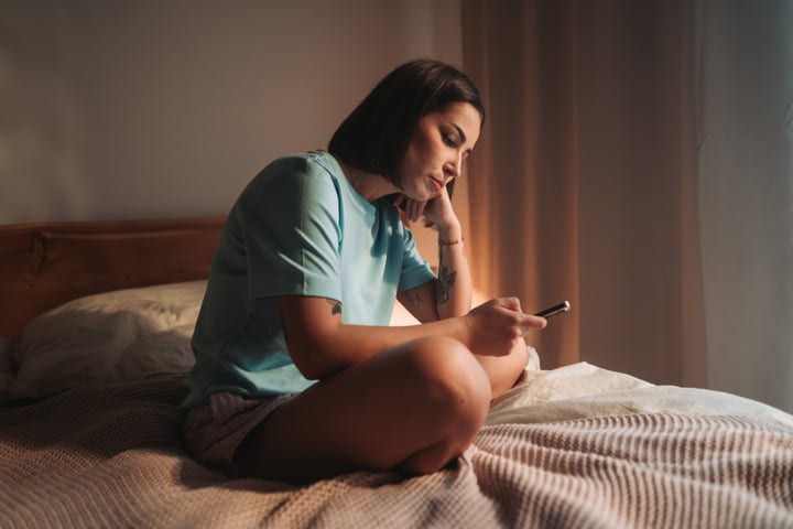 Young Woman Sits On Bed Browsing Her Smartphone In Soft Morning Light