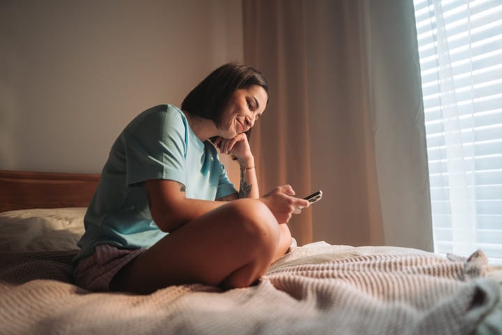 Young Woman In Bed Browsing Smartphone Content On A Sunny Morning