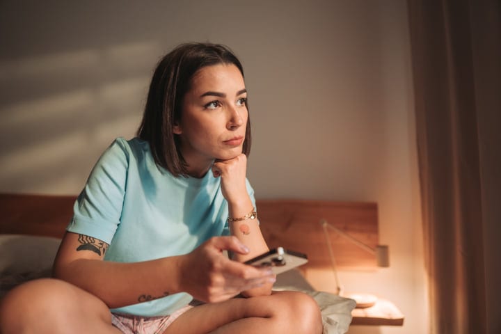 Young Woman Sits On Bed Looking Out Window Holding Phone In Morning Light