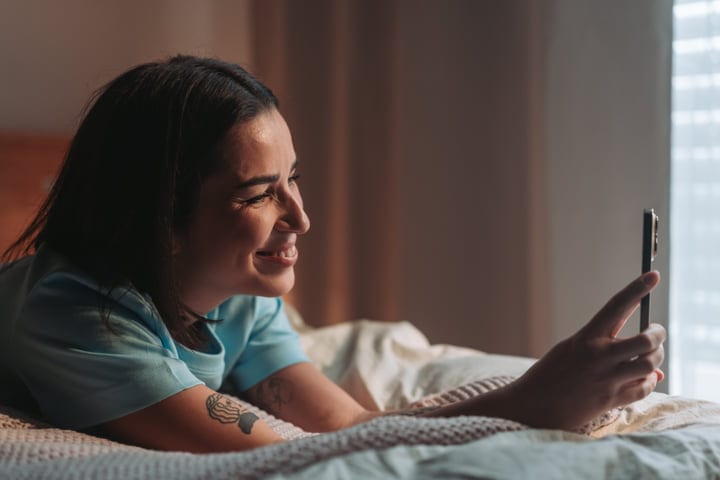 Smiling Woman Relaxes In Bed Reading On Smartphone In Morning Light