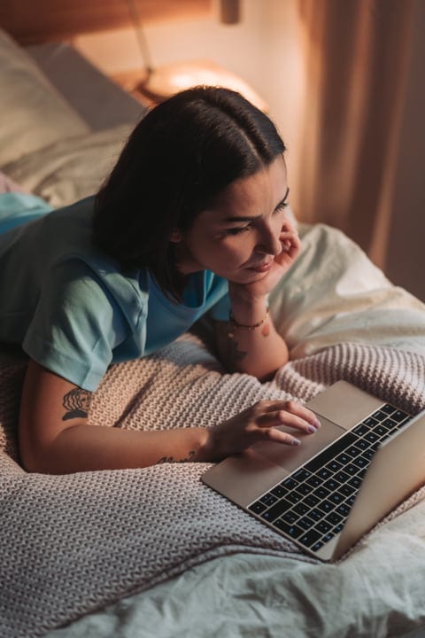 Young Woman Lying On Bed Using Laptop In Soft Morning Light