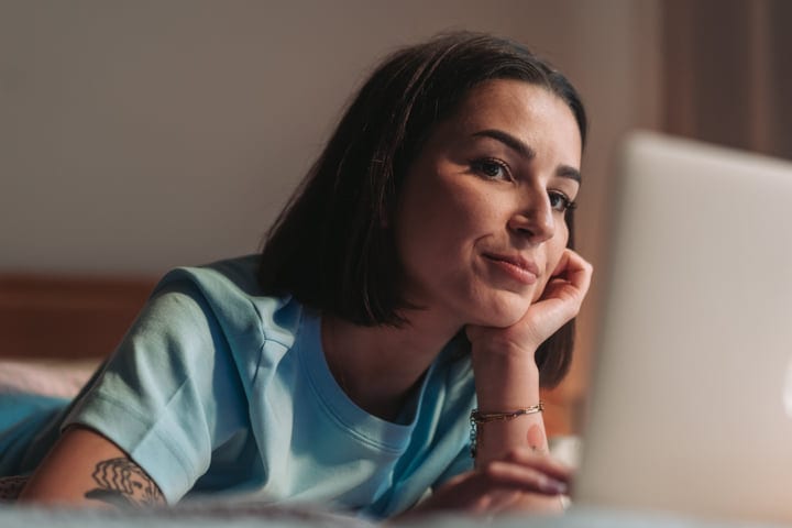 Young Woman Smiling While Using Laptop In Bed Early Morning