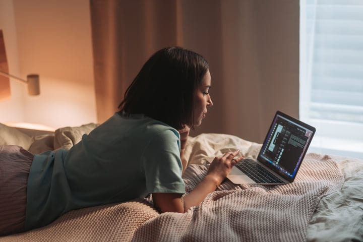 Young Woman Lying On Bed Using Laptop In Cozy Bedroom Morning Light