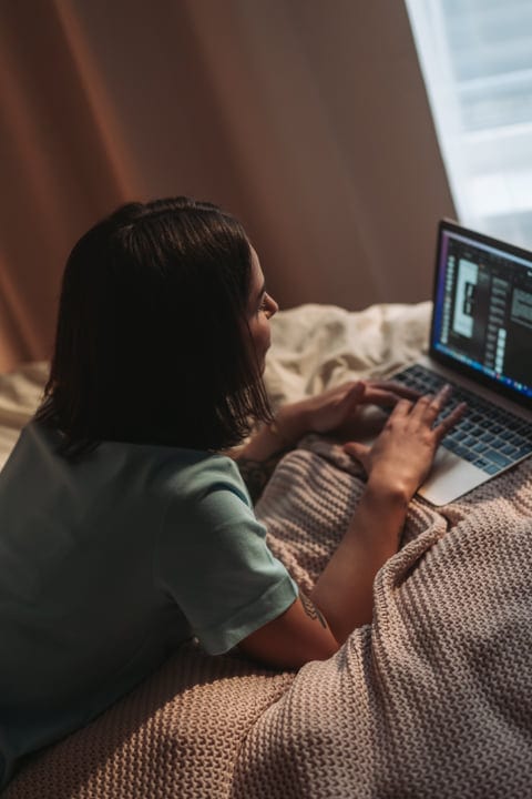 Woman Typing On Laptop In Bed With Blanket Next To Window Light