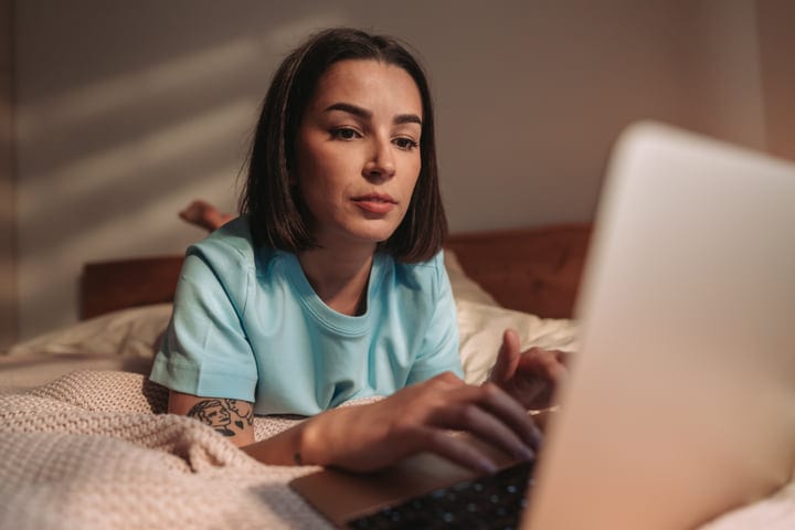 Young Woman Lying On Bed Focused On Laptop In Morning Light