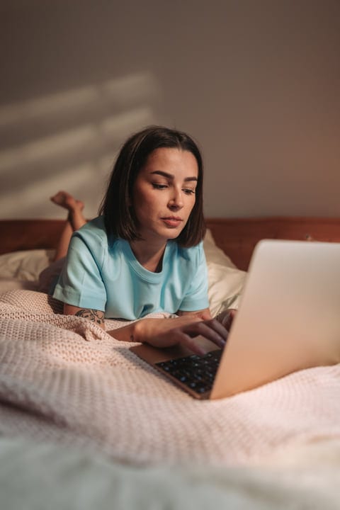 Young Woman Lying On Bed Working On Laptop In Cozy Bedroom