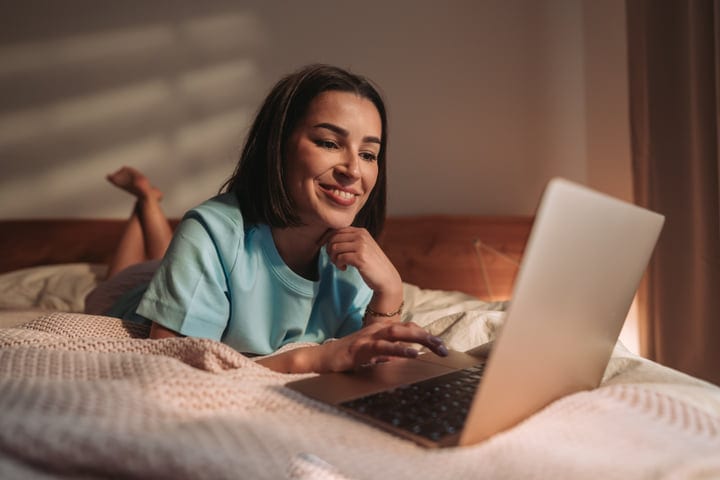 Smiling Woman On Bed Using Laptop In Cozy Bedroom Light