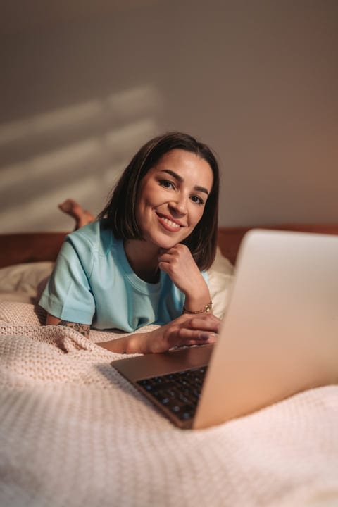 Young Woman Lying On Bed Smiling Using Laptop In Bedroom