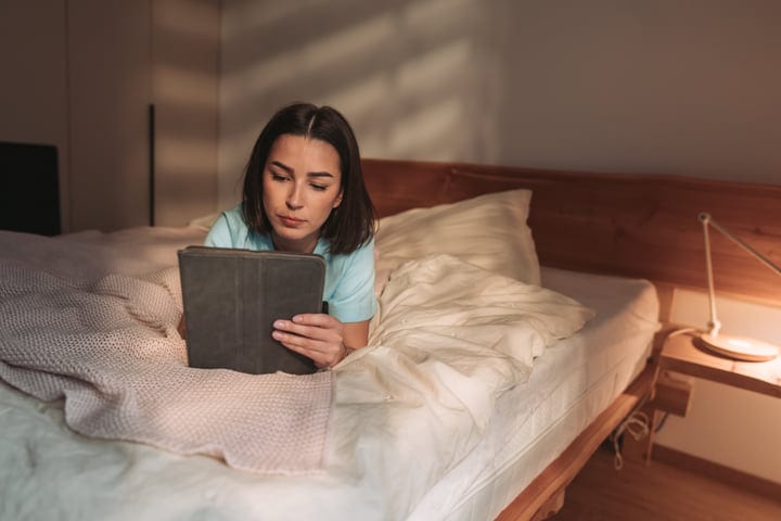 Young Woman Lying On Bed Using Her Tablet In A Cozy Bedroom