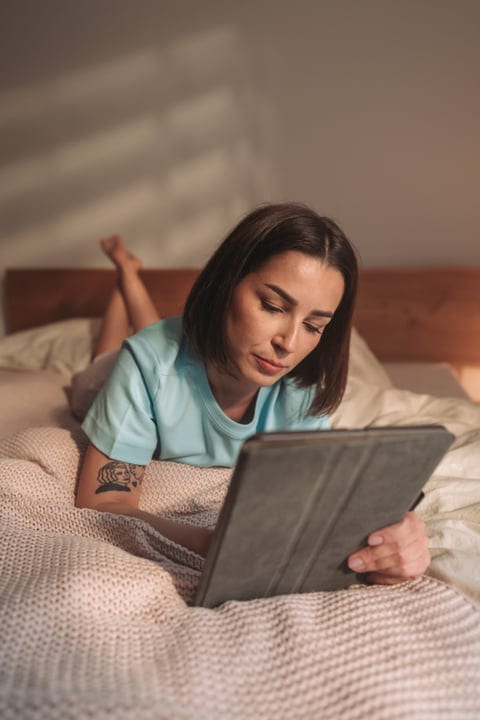Young Woman Lies On Bed Reading Tablet In Soft Morning Light
