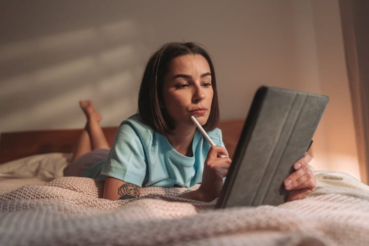 Young Woman Lying on Bed Using Digital Tablet with Stylus Indoors