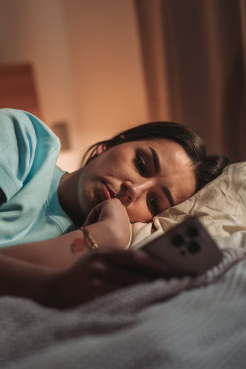 Brunette Woman In Hoodie Lying Down Browsing Smartphone In Bedroom