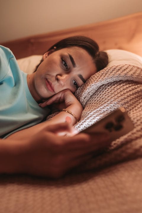 Woman Lying In Bed Using Smartphone Under Blanket During Morning