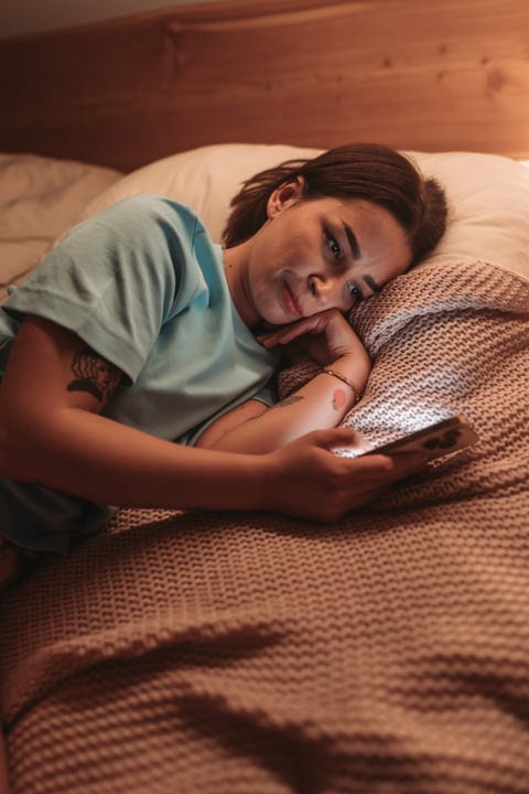 Woman Lying In Bed Browsing Her Smartphone In The Morning Light