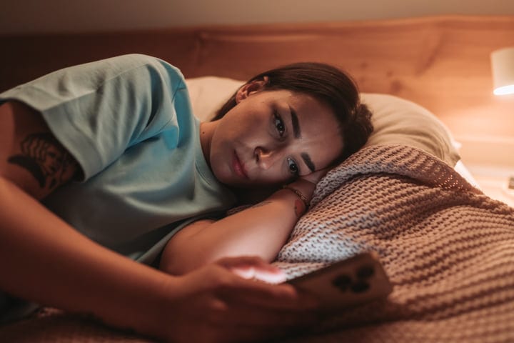 Young Woman Lying In Bed Checking Her Smartphone In A Dimly Lit Room