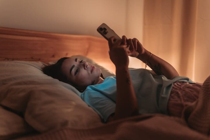 Young Woman Lying In Bed Scrolling On Smartphone In Warm Morning Light