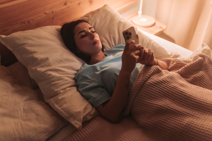 Woman Relaxing In Bed Checking Her Smartphone In Warm Lit Room