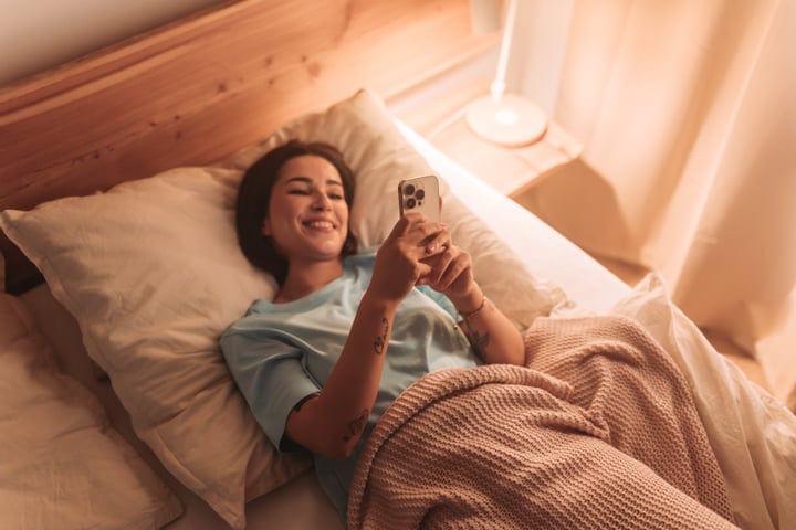 Happy Woman Lying In Bed And Using Her Smartphone In Morning