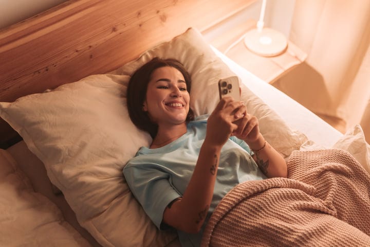 Young Woman Lying In Bed Smiling And Looking At Her Smartphone