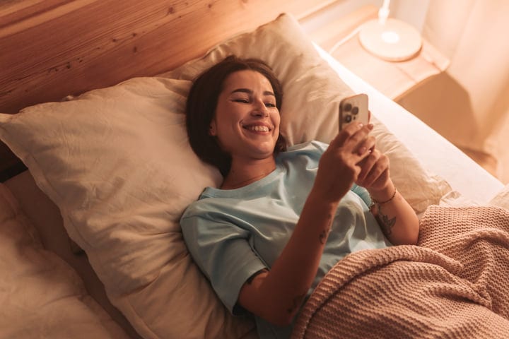 Young Woman Smiles Using Phone In Bed With Morning Sunlight