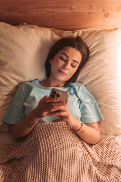 Young Woman In Bed Browsing Smartphone In Soft Morning Glow