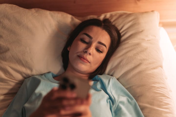 Young Woman Lying In Bed Scrolling Smartphone In Dim Morning Light