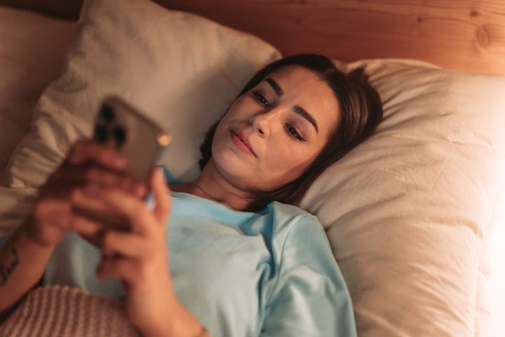 Young Woman Waking Up In Bed And Checking Her Smartphone