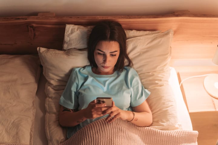 Brunette Woman Reading Messages On Her Smartphone From Bed In The Morning