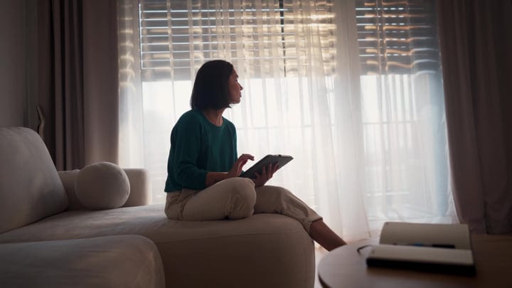 Woman Sitting On Sofa Using Tablet By Window In Bright Morning Light