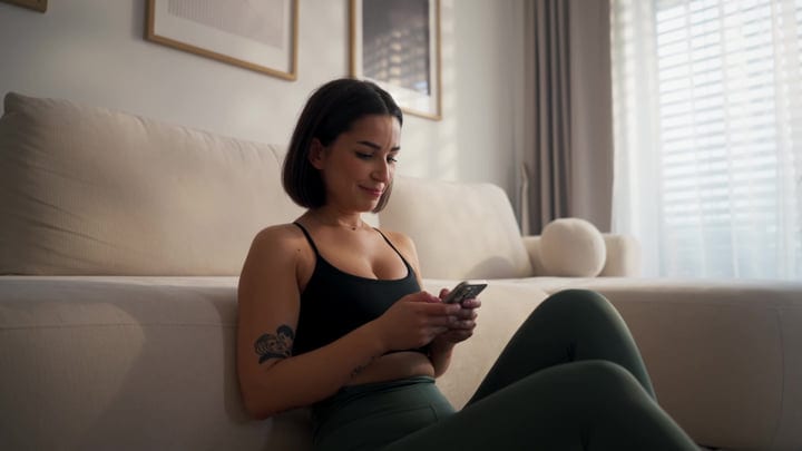 Woman Smiling, Using Phone On Floor Beside Sofa In Living Room