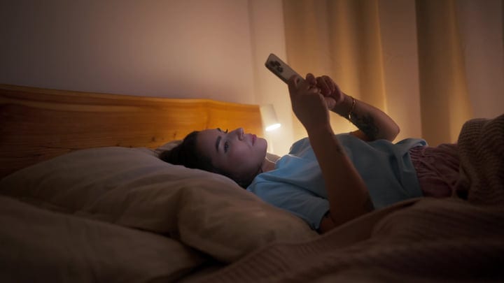 Young Woman Lying In Bed Using Smartphone At Night In Dimly Lit Bedroom