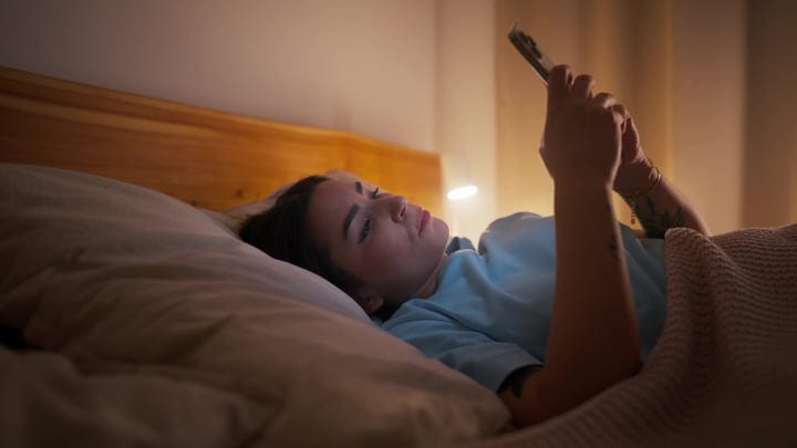 Young Woman Lying In Bed Scrolling On Smartphone In Dark Bedroom