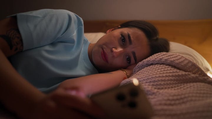 Young Woman Lying In Bed Scrolling On Smartphone In Dim Morning Light