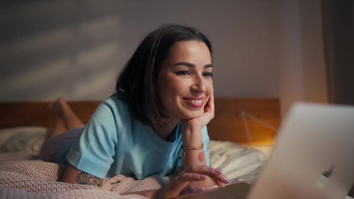 Content Young Woman Lying Comfortably On Her Bed Using A Laptop Computer