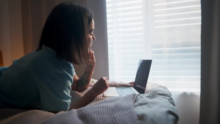 Young Woman Relaxing On Bed While Watching Laptop In Her Bedroom