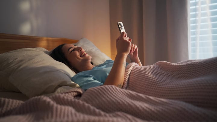 Young Woman Laughing While Using Smartphone In Bed At Morning