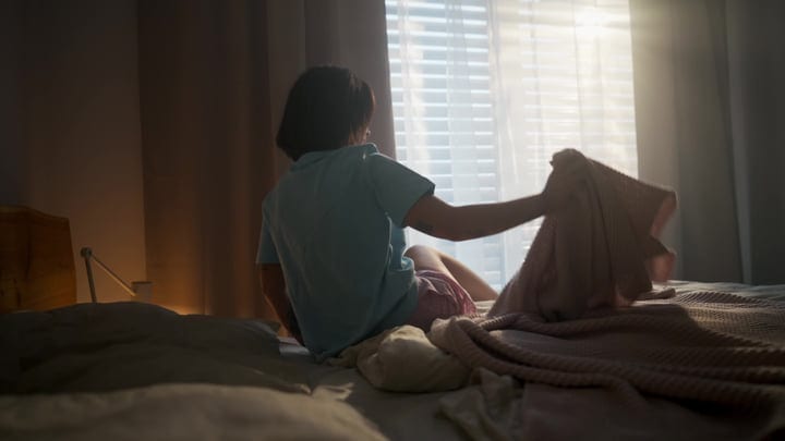 Woman Sits On Bed Reaching For Blanket As Morning Sunlight Streams Through Window