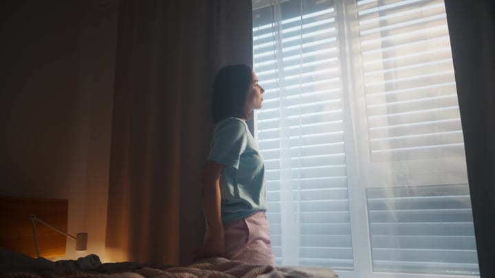 Woman Looks Out Bedroom Window At Bright Morning Light Through Blinds