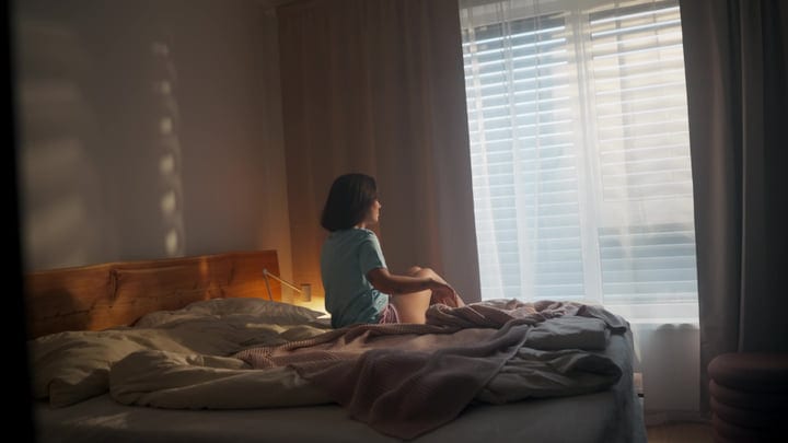 Woman Sits On Bed Looking Out Bedroom Window In Morning