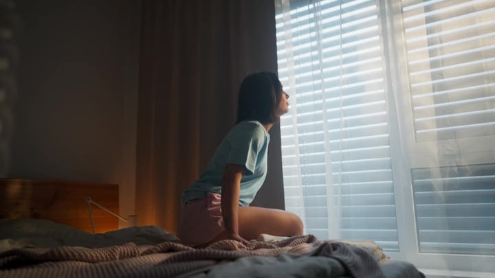 Young Woman Sits On Bed Looking Out Window Through Blinds At Early Morning Light