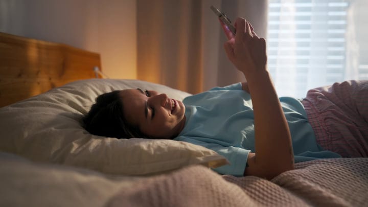 Young Woman Lying In Bed Smiling At Her Smartphone