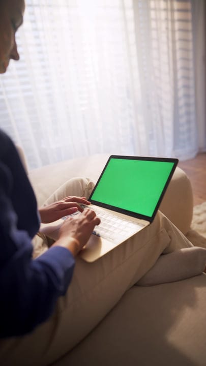 Woman Typing On Laptop With Green Screen, Sitting On A Sofa By A Sunlit Window