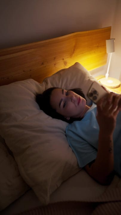 Young Woman Relaxing In Bed Scrolling On Her Smartphone At Night